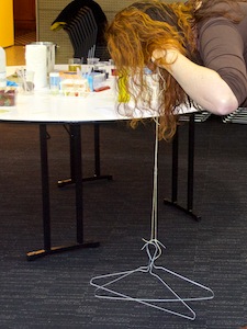 a woman (mostly her hair) bending over, fingers in ears, has two coathangers attached by string to her fingers